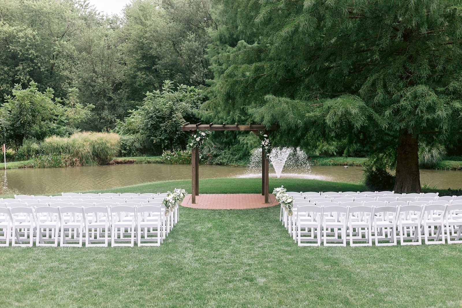 image shows rows of white chairs and an arch decorated with greenery, all set up for an outdoor wedding by the pond at historic acres of hershey