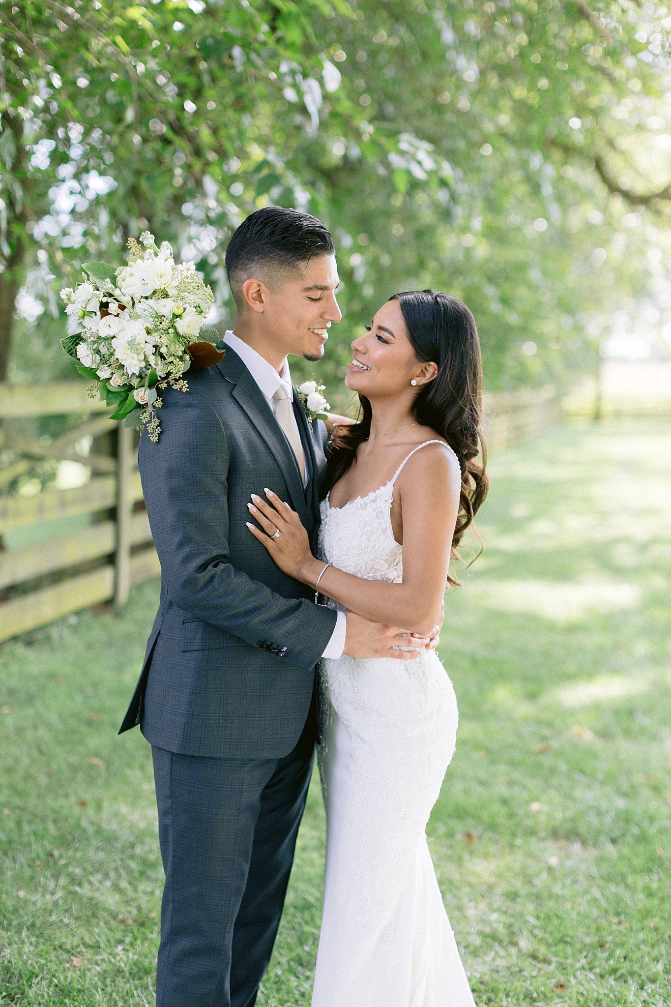 York college alumni Ailin and Anthony hold each other close for their wedding portraits with Tom and Em Weddings. He is wearing a grey fitted suit, holding his new wife by her waist. She is wearing a fitted white lace dress with detailed lace bodice, a long train, and thin straps. They are standing among the trees at their wedding venue.